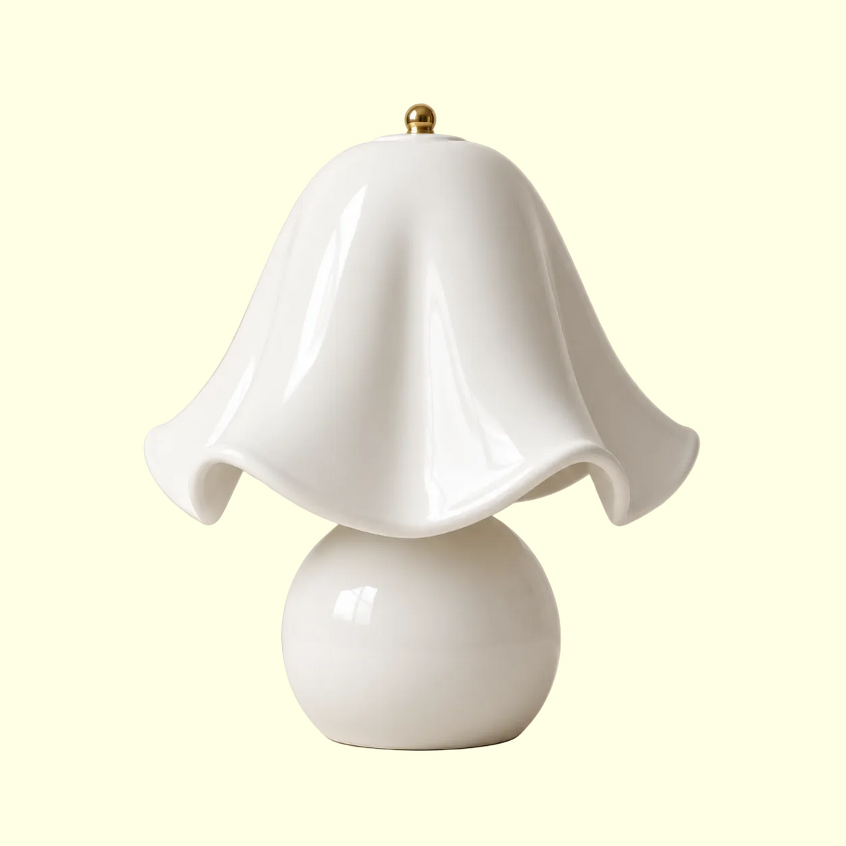 A glossy white ceramic table lamp with a wavy, bell-shaped shade and a round base, topped with a small gold finial, set against a solid beige background.