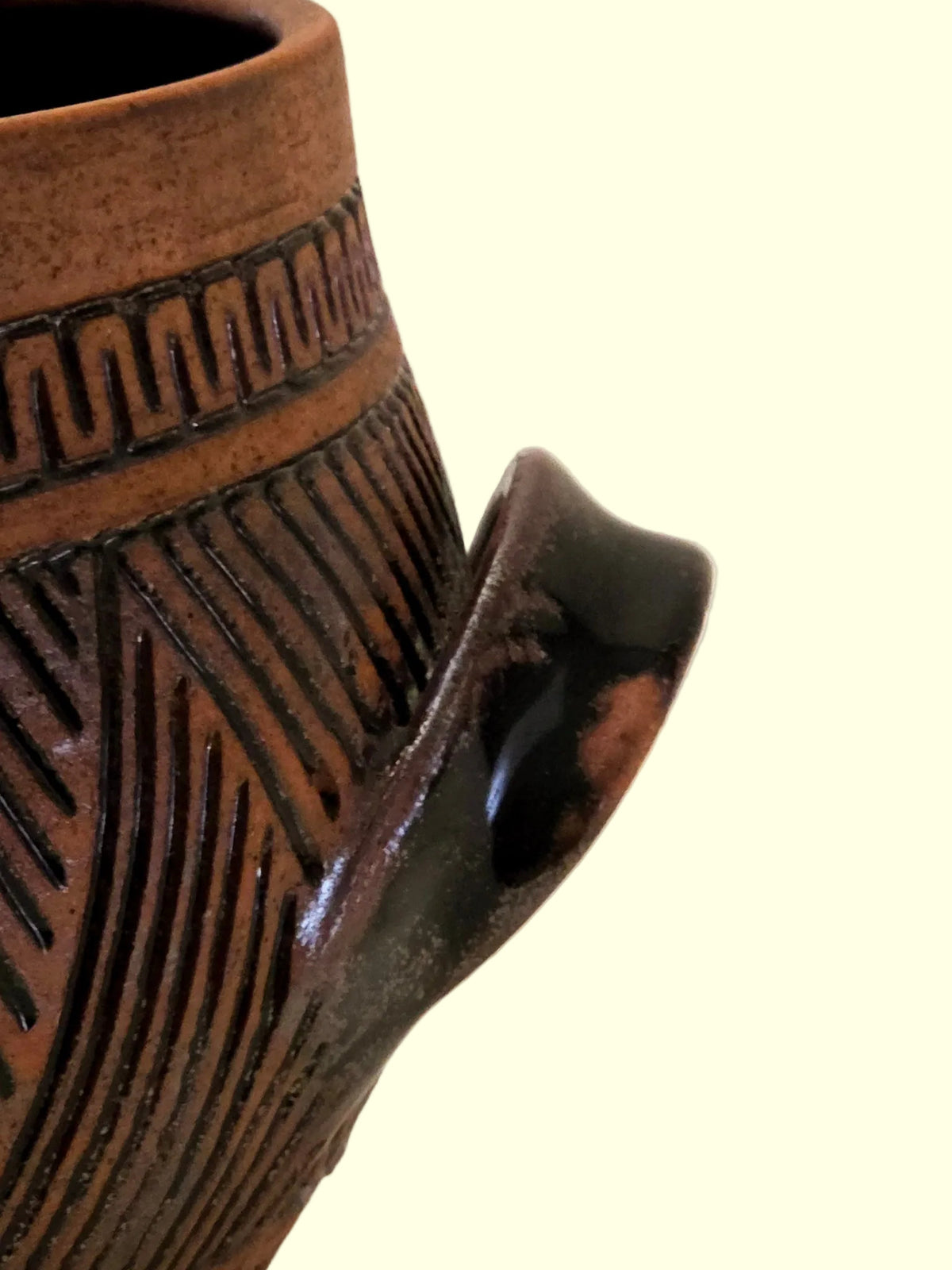 Close-up of a handmade terracotta clay pot with carved geometric patterns and a dark glazed handle, set against a plain off-white background.