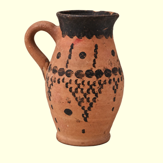 A terracotta-colored ceramic pitcher with a black hand-painted handle, rim, and geometric dot patterns on the body, set against a plain light beige background.
