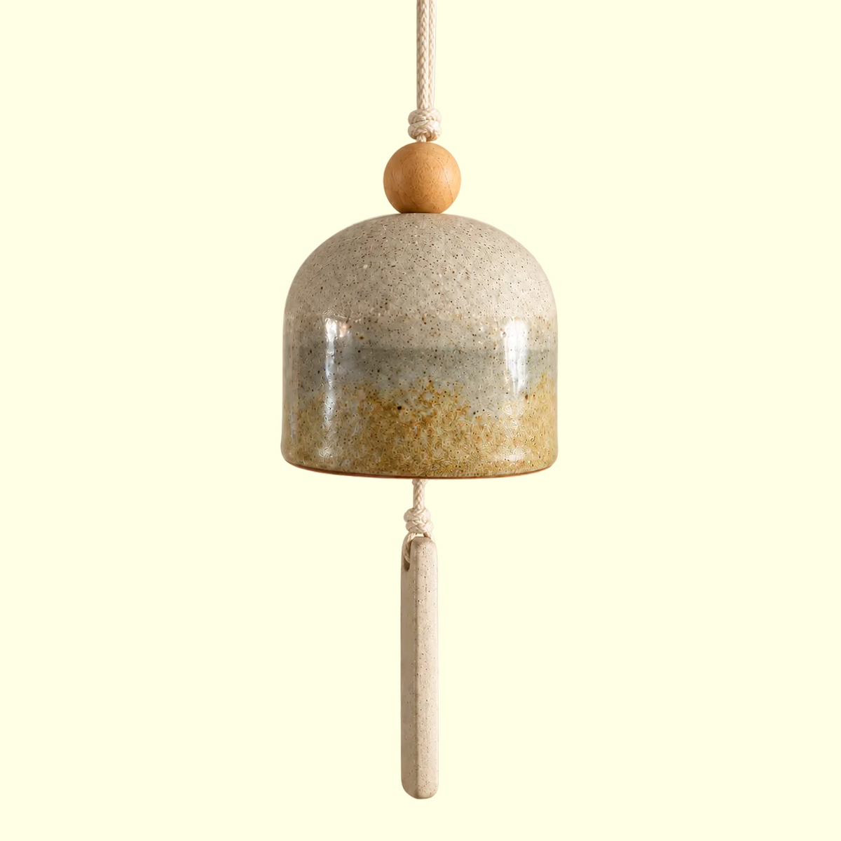 A hanging ceramic wind chime bell with a speckled beige and gray glaze, a natural wood bead on top, and a long matching ceramic tassel, shown against a solid warm beige background.