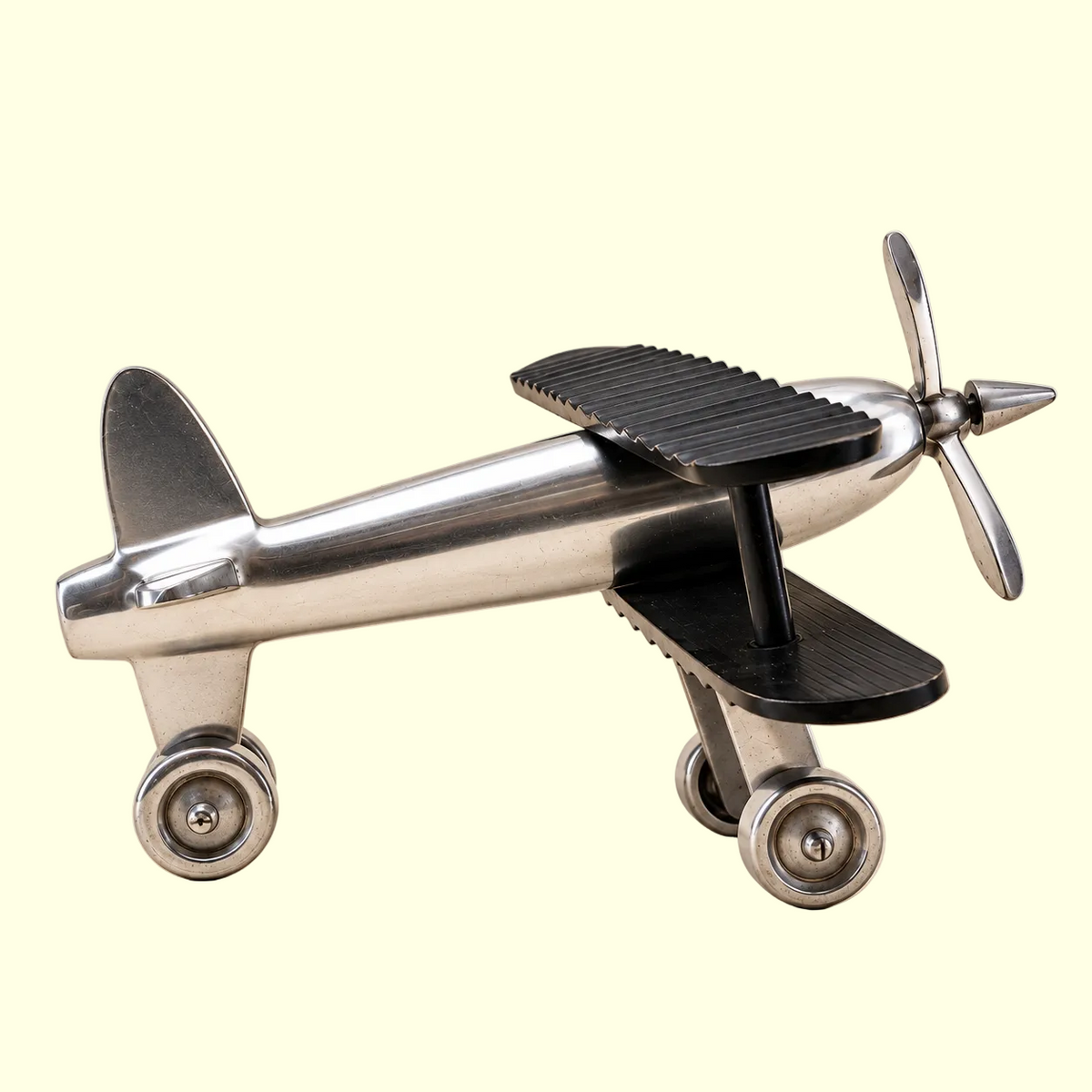A polished stainless steel toy or decorative biplane with black textured wing panels and silver propeller blades, photographed on a plain warm beige background.