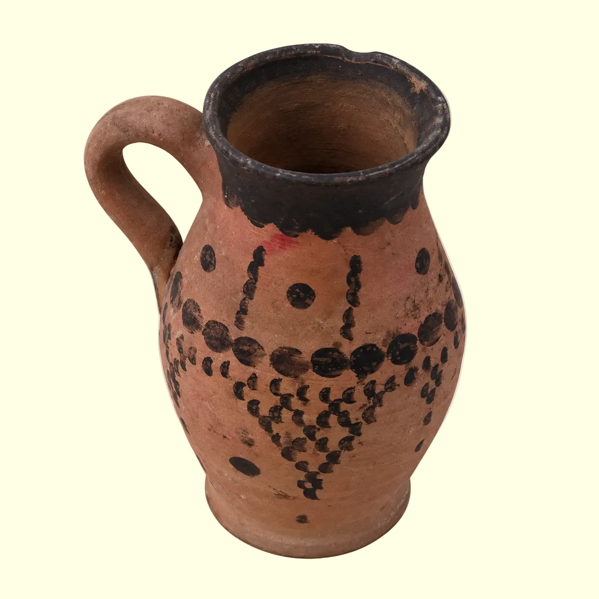 A terracotta-colored ceramic pitcher with a dark brown to black painted rim and decorative geometric dot patterns on the body, set against a plain cream background.