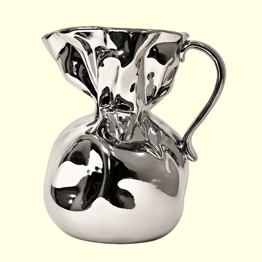 A polished metallic pitcher with a sculptural, fluid, organic shape and a curved handle, shown on a plain light beige background.