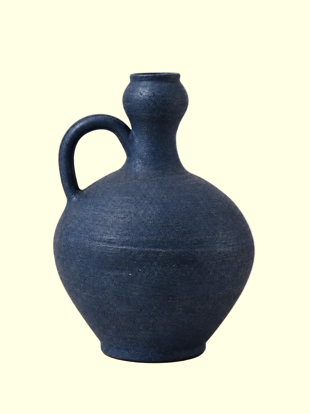 A dark blue textured ceramic jug with a rounded body, a side loop handle, and a narrow neck, set against a solid light beige background.