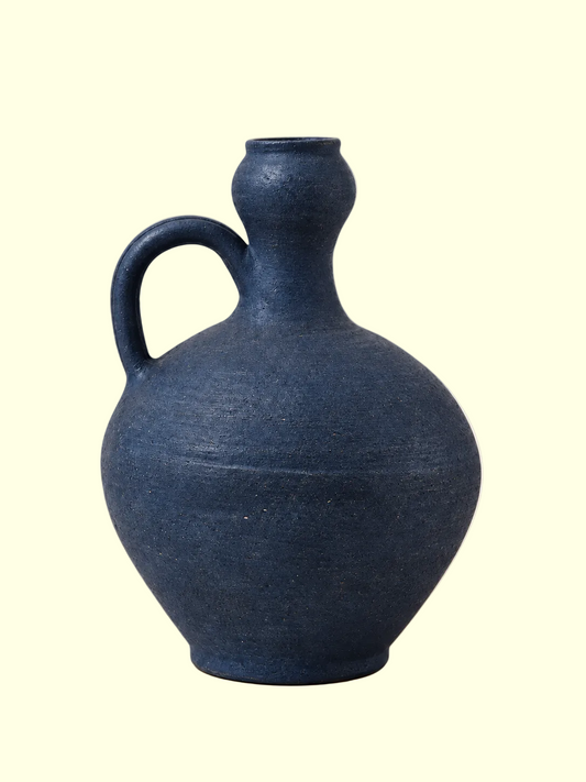 A dark blue textured ceramic jug with a rounded body, a side loop handle, and a narrow neck, set against a solid light beige background.