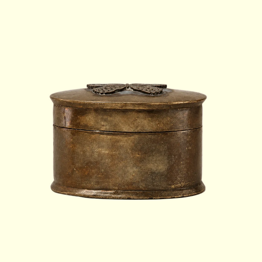 An antique oval metal box with a distressed bronze finish, featuring a butterfly-shaped metal clasp on the lid, set against a plain off-white background.