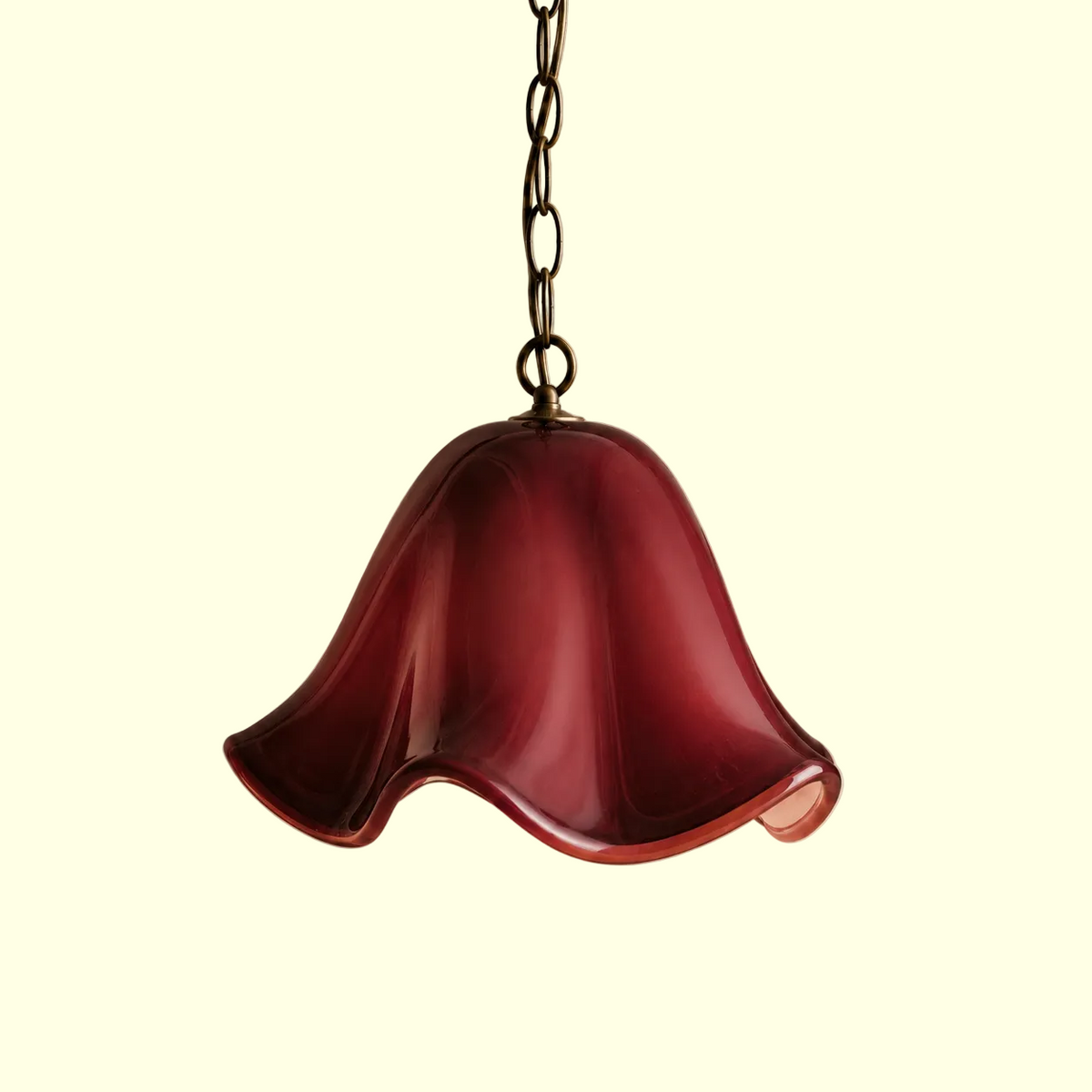 A hanging pendant light with a translucent bell-shaped shade in deep burgundy glass, suspended from a dark metal chain against a warm beige textured wall.