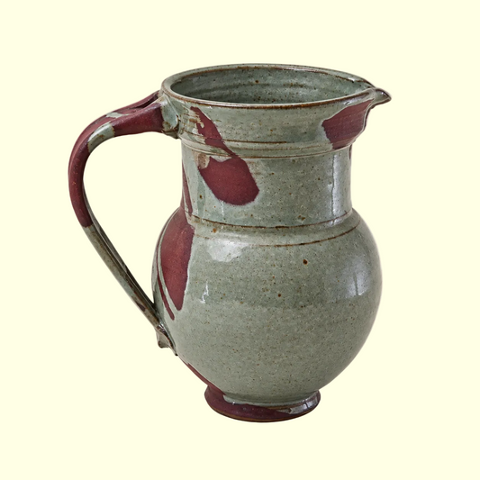 A handmade ceramic pitcher with a rounded body, curved handle, and small pouring spout, featuring a speckled sage green glaze with dark reddish-brown accents, set against a solid light beige background.