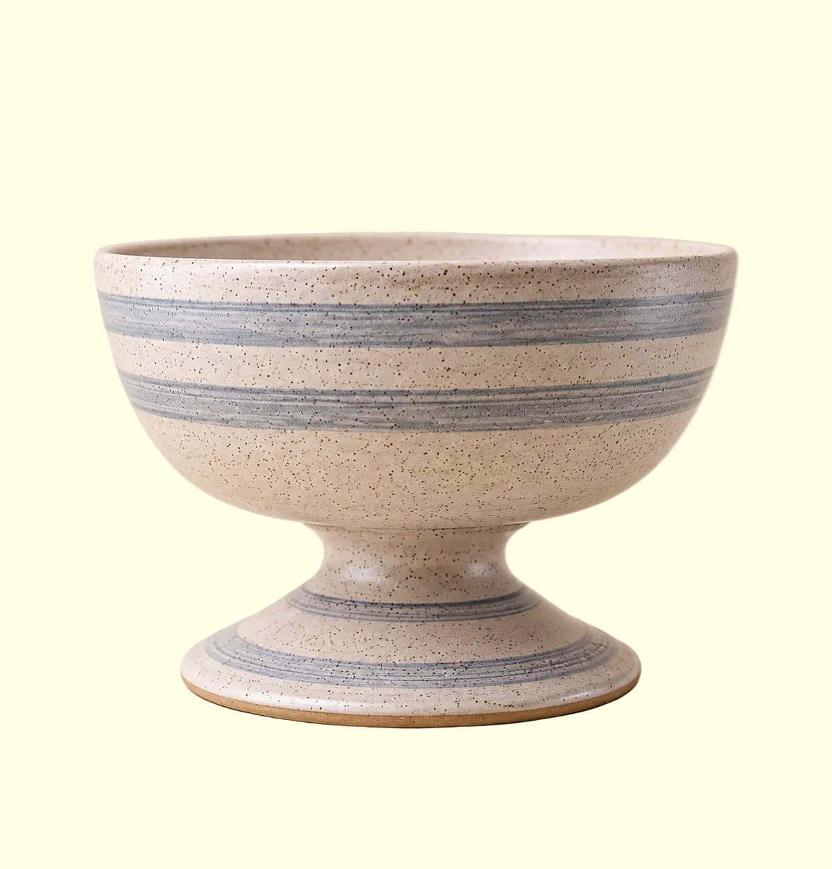 A handmade ceramic pedestal bowl with a speckled beige glaze and horizontal blue stripes, set against a solid warm beige background.
