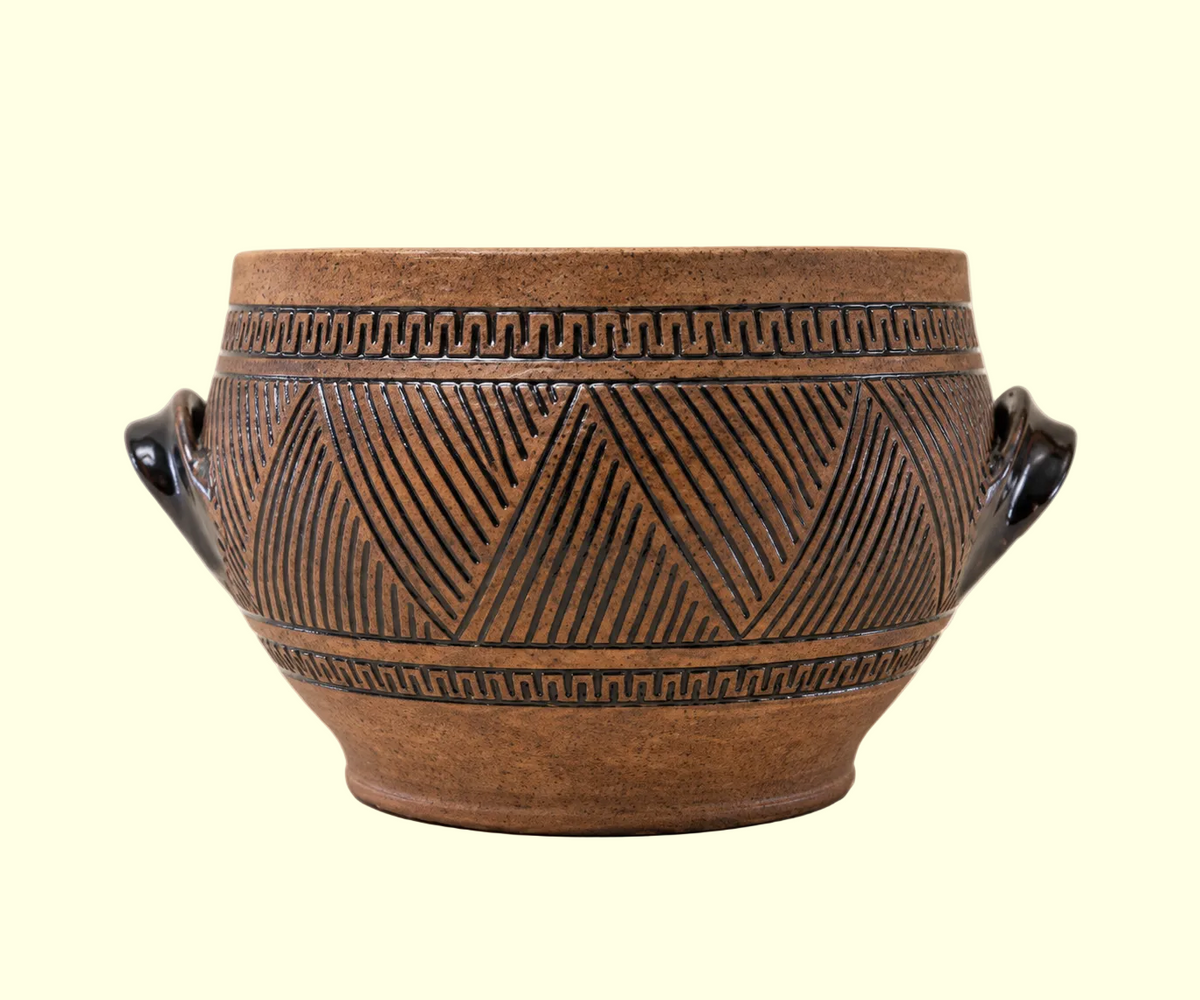A brown ceramic pot with carved geometric patterns and two dark handles, photographed against a plain light beige background.