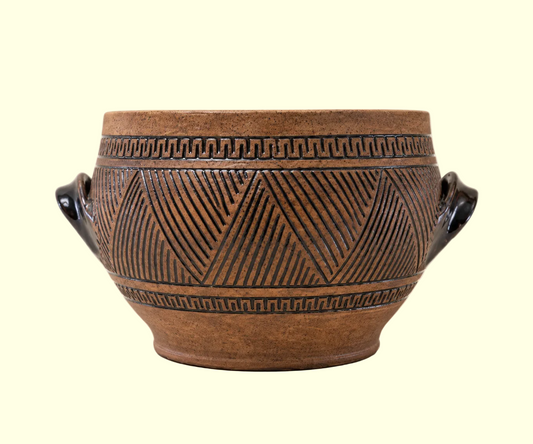 A brown ceramic pot with carved geometric patterns and two dark handles, photographed against a plain light beige background.
