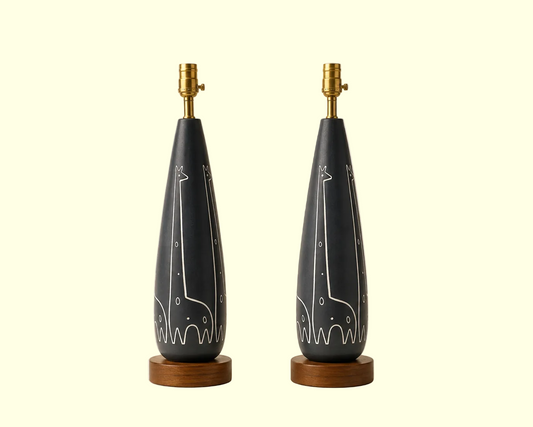 Two matching raymor table lamps with dark charcoal gray teardrop-shaped ceramic bases featuring white giraffe line drawings, mounted on round wooden bases with brass lamp sockets, set against a warm beige background.